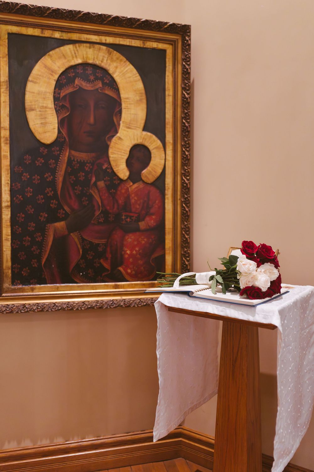 Black Madonna icon painting in gilded frame beside floral bouquet on white linen table in church.