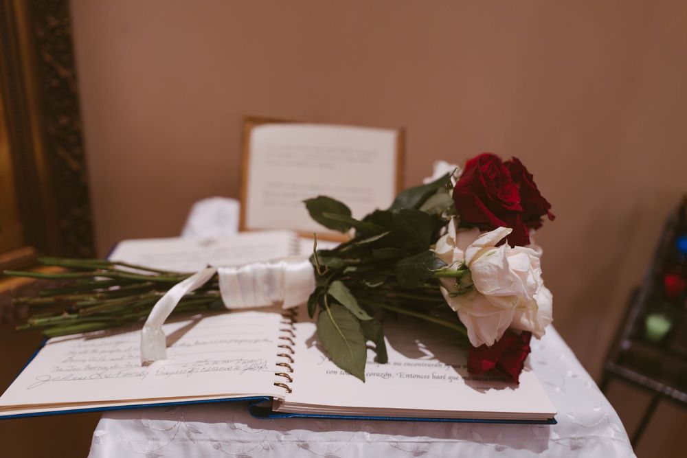 Red and white rose bouquet with white ribbon resting on an open wedding guest book on a lace-covered table.