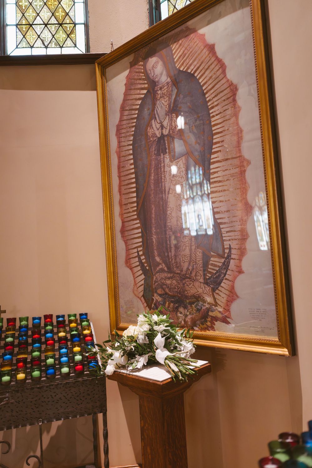 Our Lady of Guadalupe framed painting displayed on church wall with white floral arrangement and colorful votive candles.