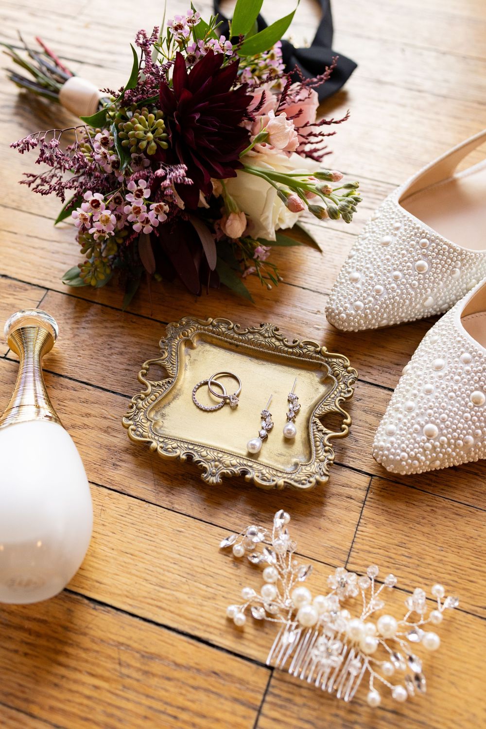 Bridal flat lay with pearl shoes, gold ring dish, diamond earrings, wedding rings, hair comb, perfume, and floral bouquet.