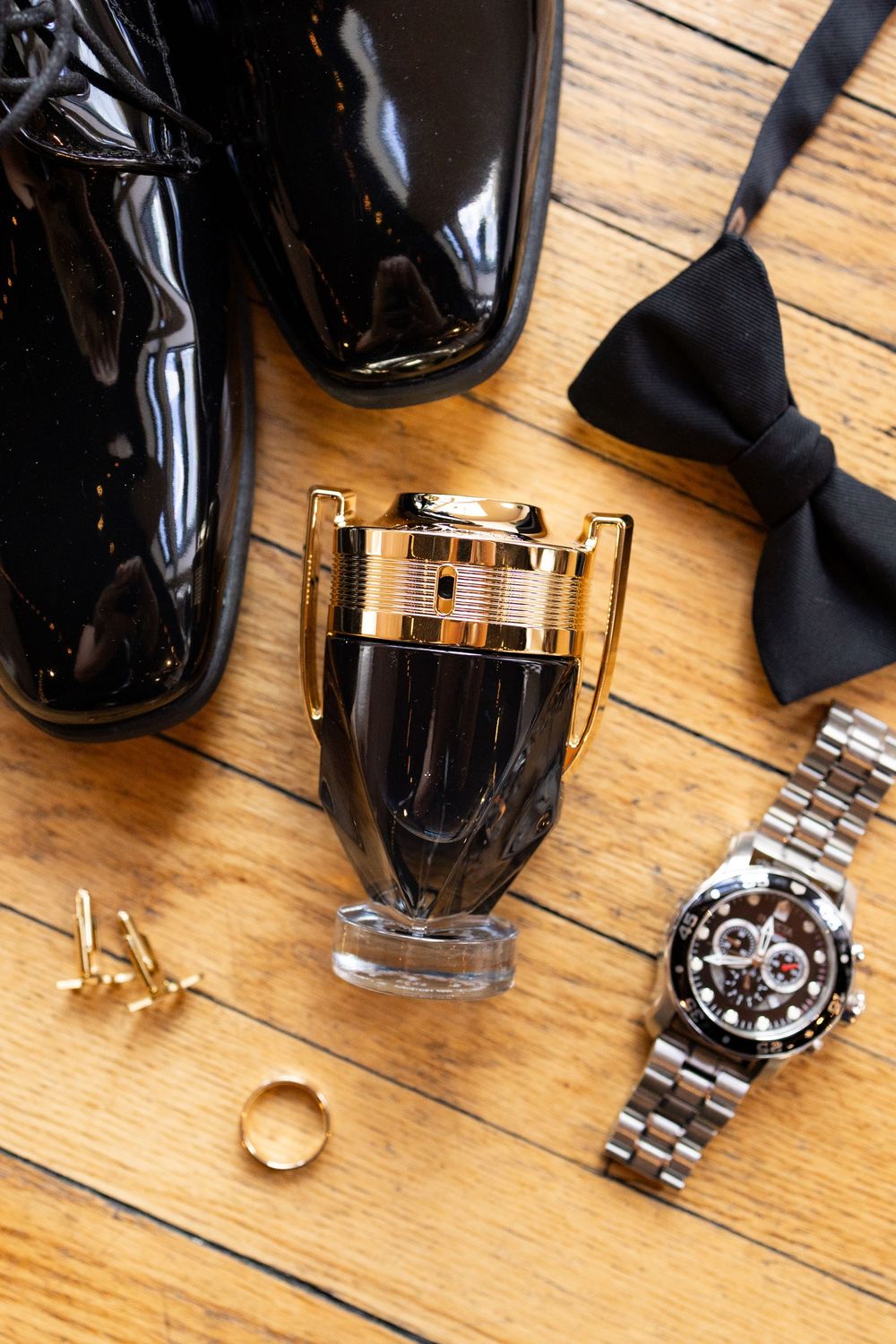 Men's accessories flat lay featuring patent leather shoes, bow tie, Rolex watch, cologne, gold ring, and cufflinks on wood.