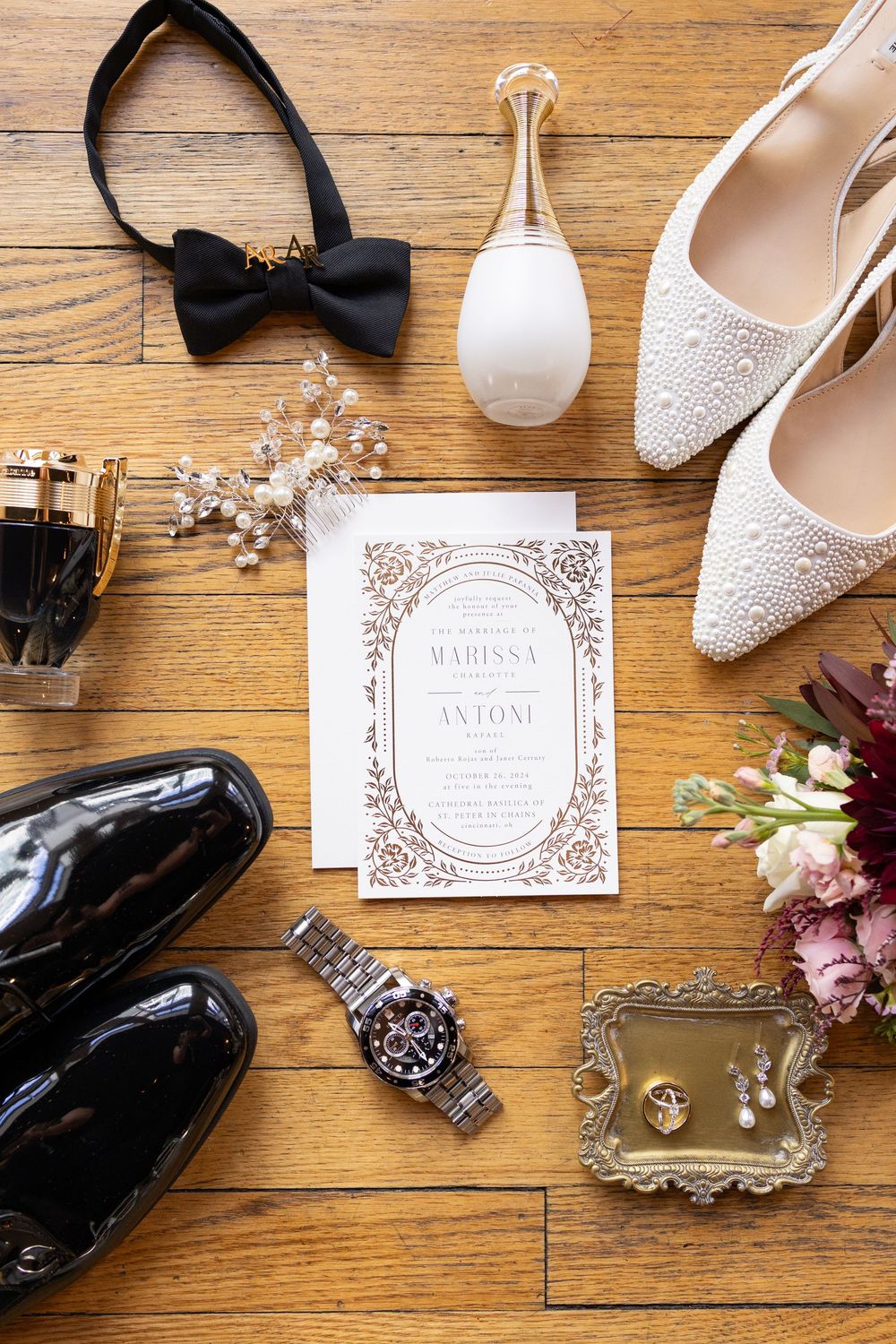 Wedding flat lay with invitation, pearl heels, bow tie, Rolex watch, floral accessories, and black patent shoes on wood floor.