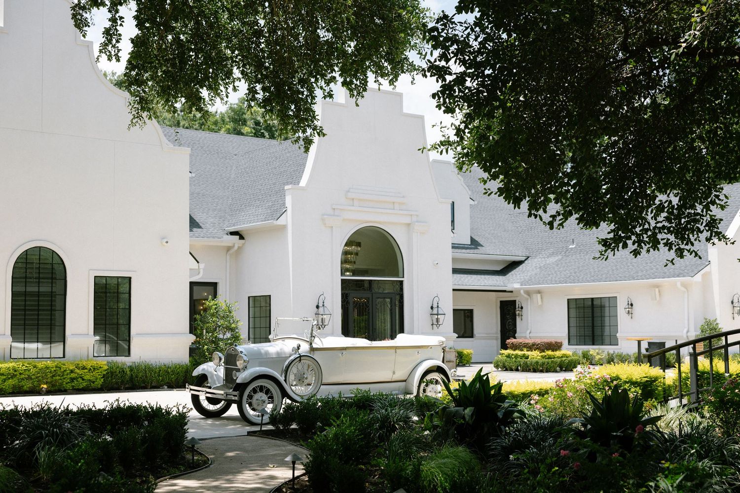 Vintage white classic car parked in front of an elegant white Cape Dutch-style mansion surrounded by lush greenery.