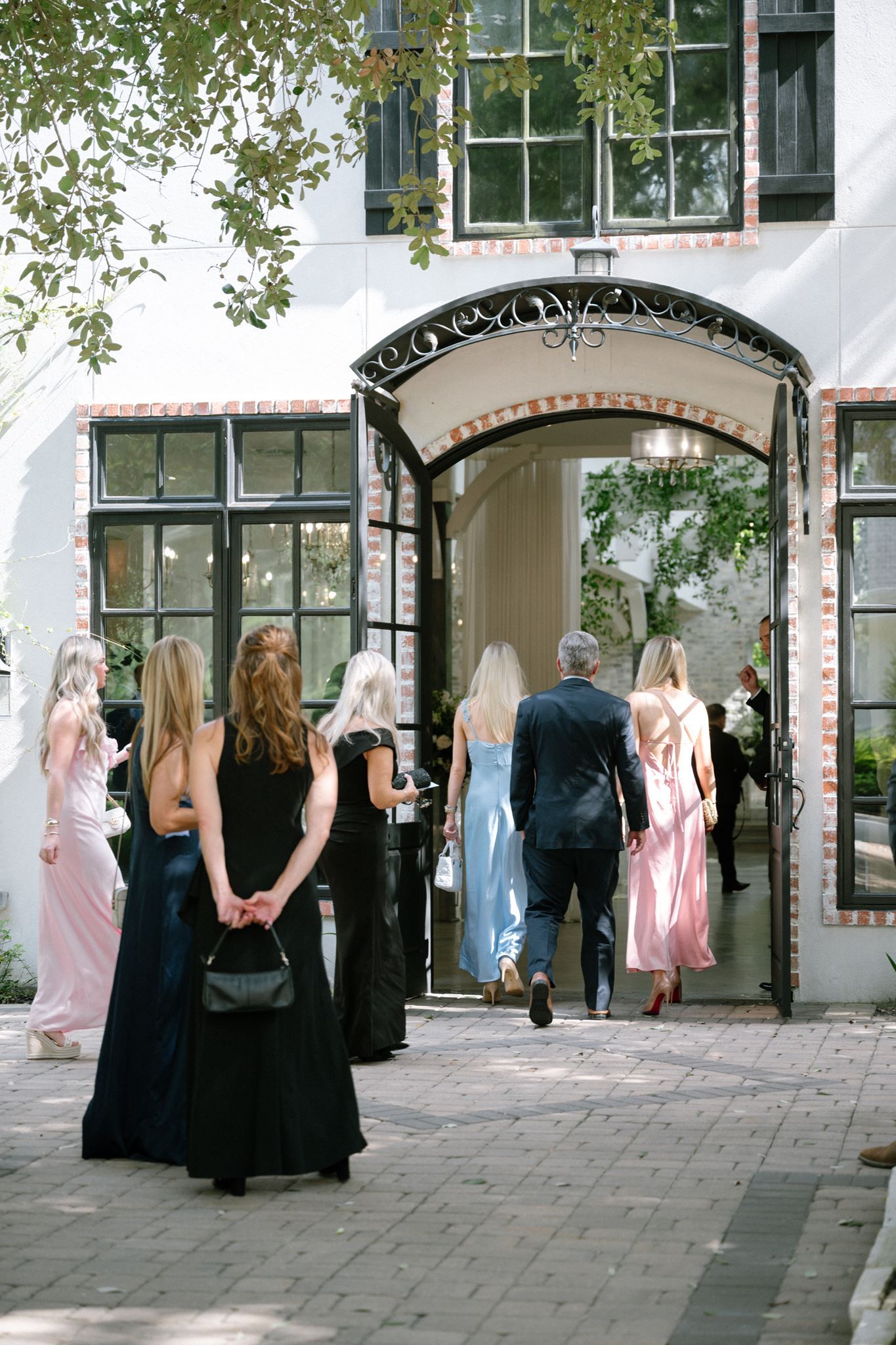 Elegantly dressed guests in formal gowns entering a white venue through ornate iron archway doors.