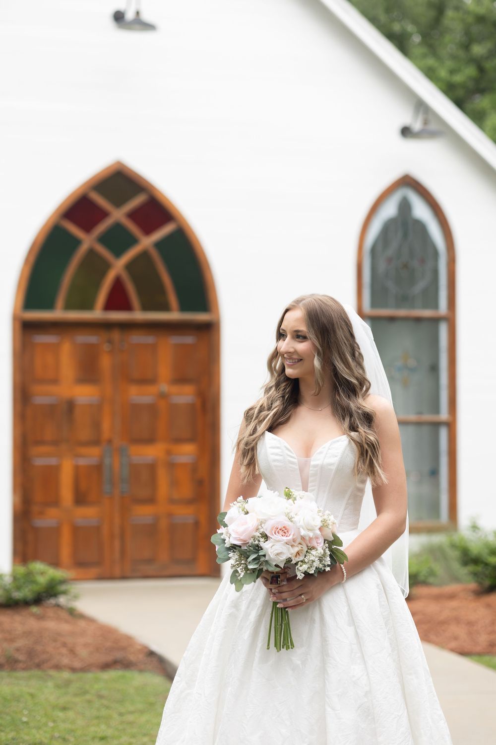 Bride in white lace gown holding pink bouquet smiles outside a white chapel with stained glass windows.