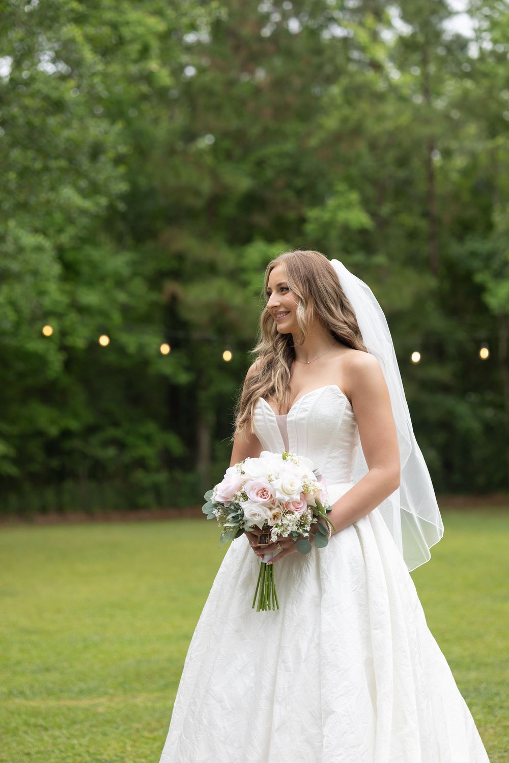 Bride in strapless lace wedding gown and veil holds pink bouquet outdoors on green lawn with string lights.