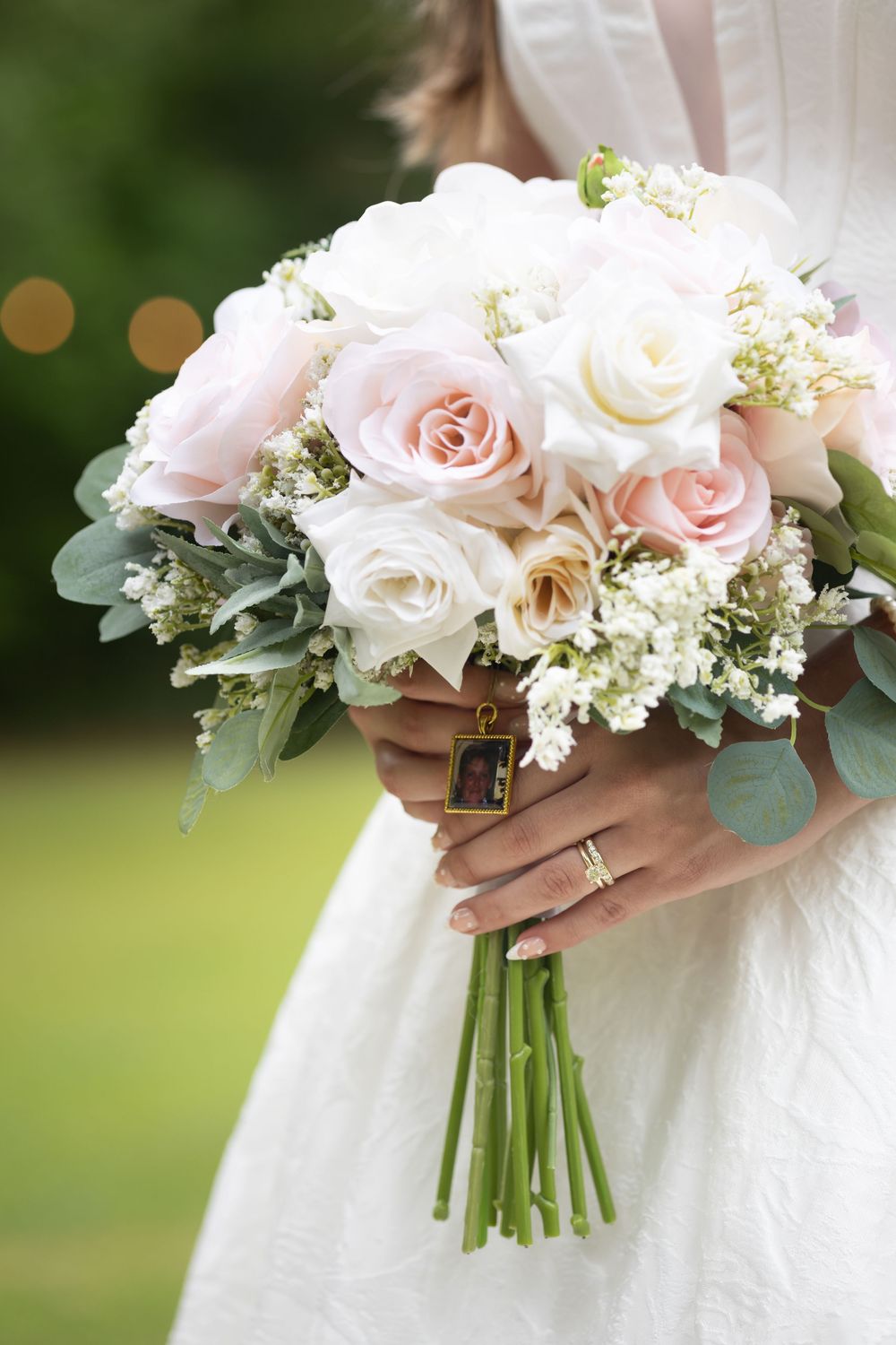 Bride holding a wedding bouquet of white and blush pink roses with eucalyptus and a small gold photo charm.