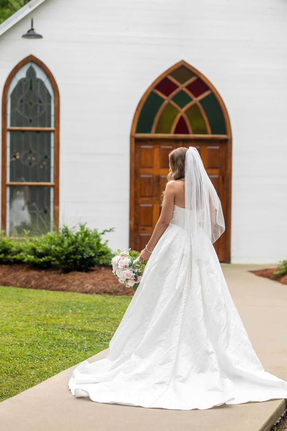 Bride in white gown and veil holds bouquet outside white chapel with stained glass windows and wooden doors.