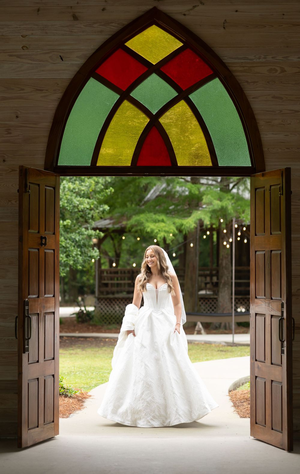 Bride in white gown standing in open chapel doors with colorful stained glass arch at outdoor wedding venue.