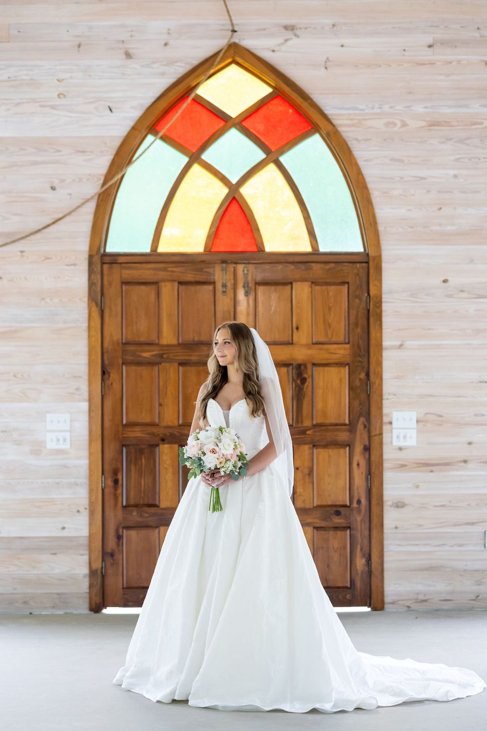 Bride in white gown with veil holds bouquet before ornate wooden doors with colorful stained glass window.