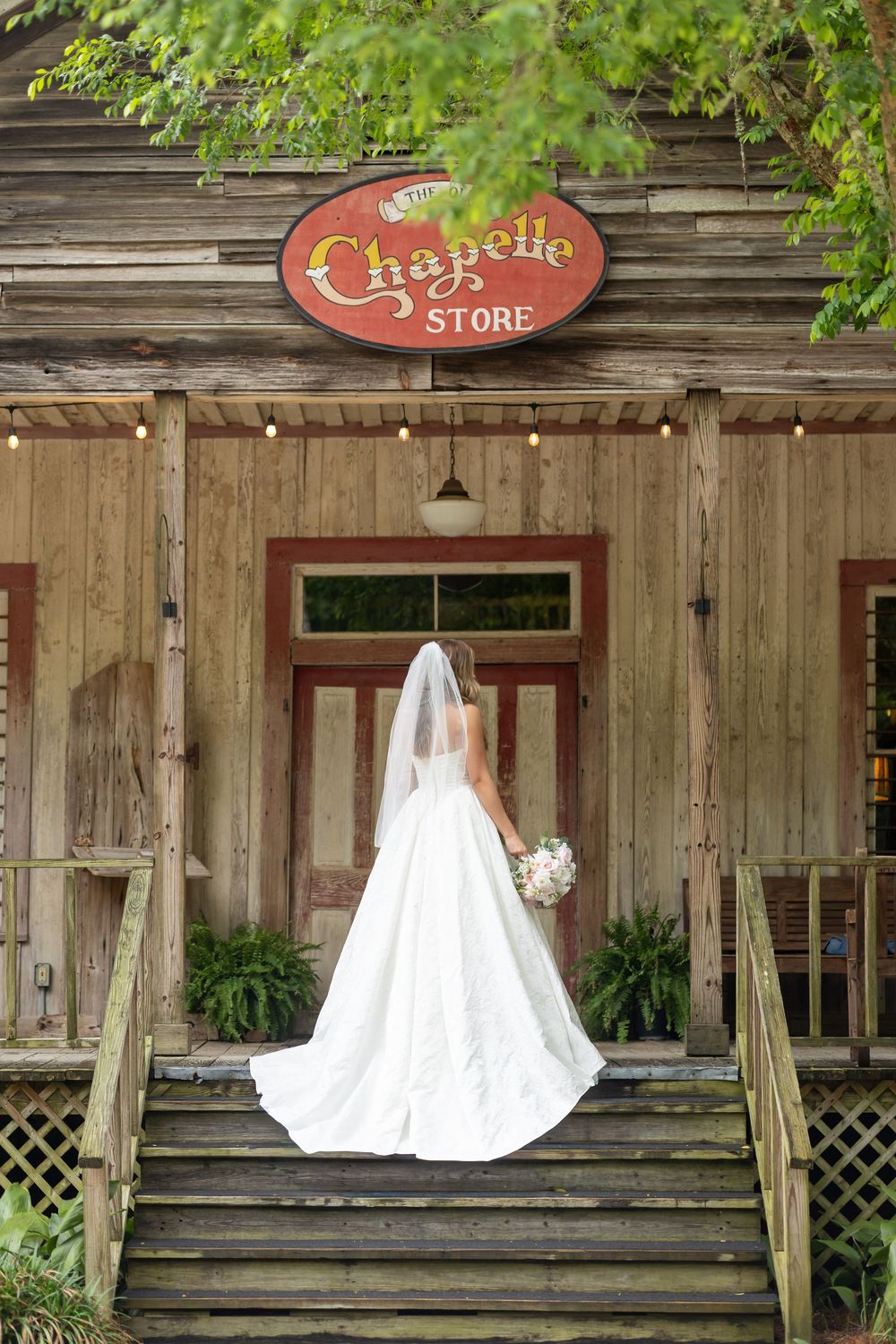Bride in white gown and veil stands on steps of rustic Chapelle Store building holding bouquet.