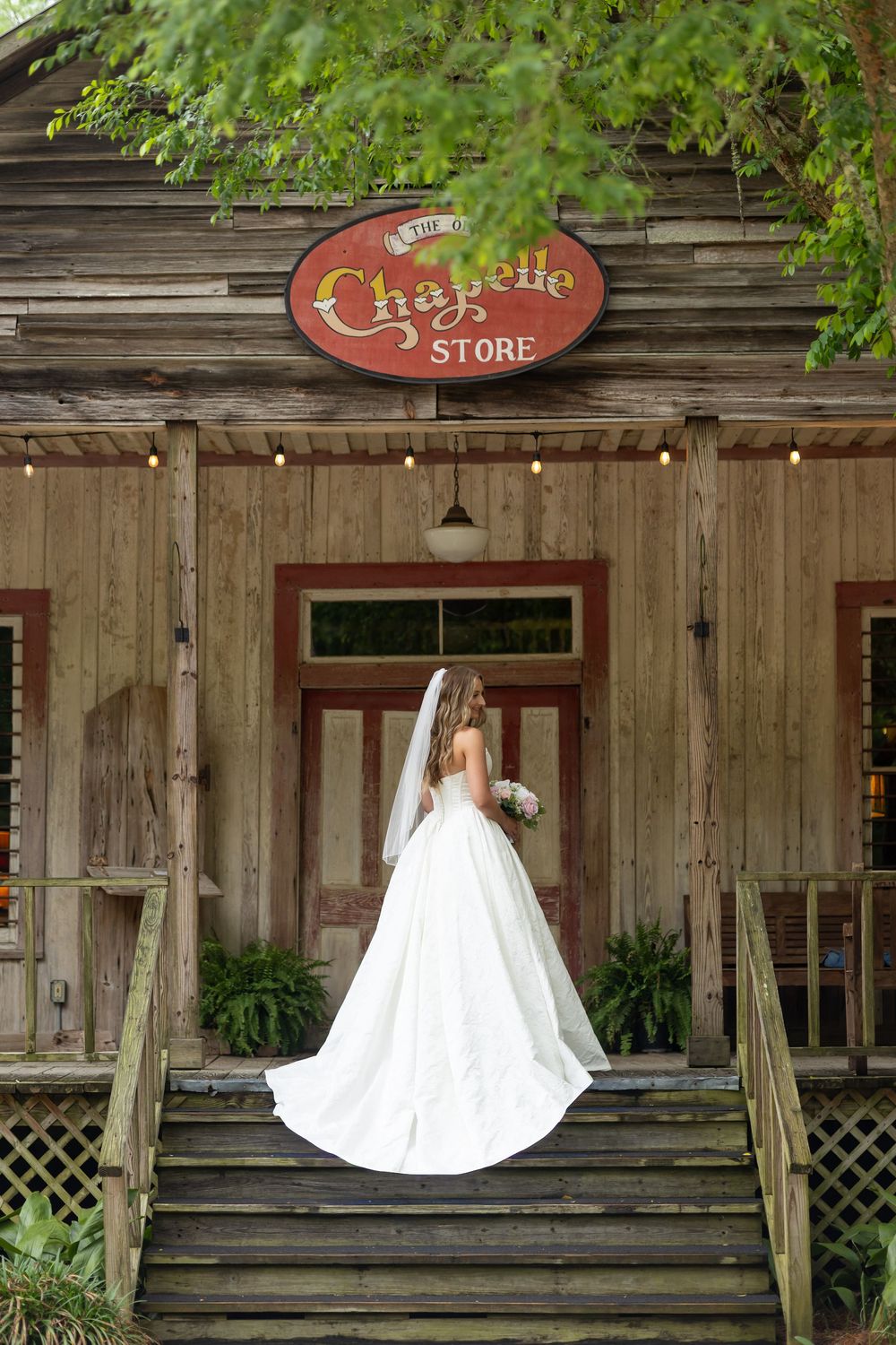 Bride in white ball gown and veil stands on steps of rustic Chapelle Store wedding venue.