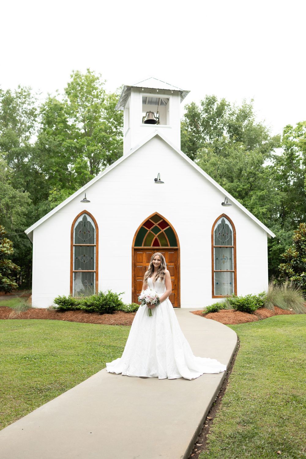 Bride in white ball gown poses before charming white chapel with arched windows and bell tower.