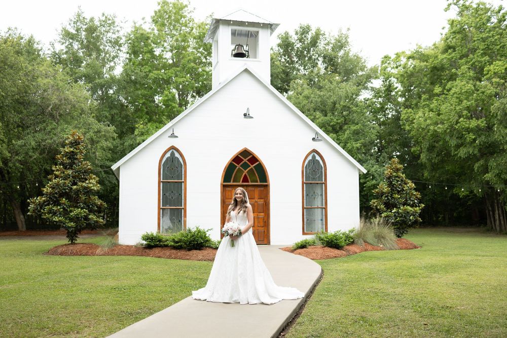 Bride in white gown poses before a charming white chapel with arched windows and wooden doors on a green lawn.