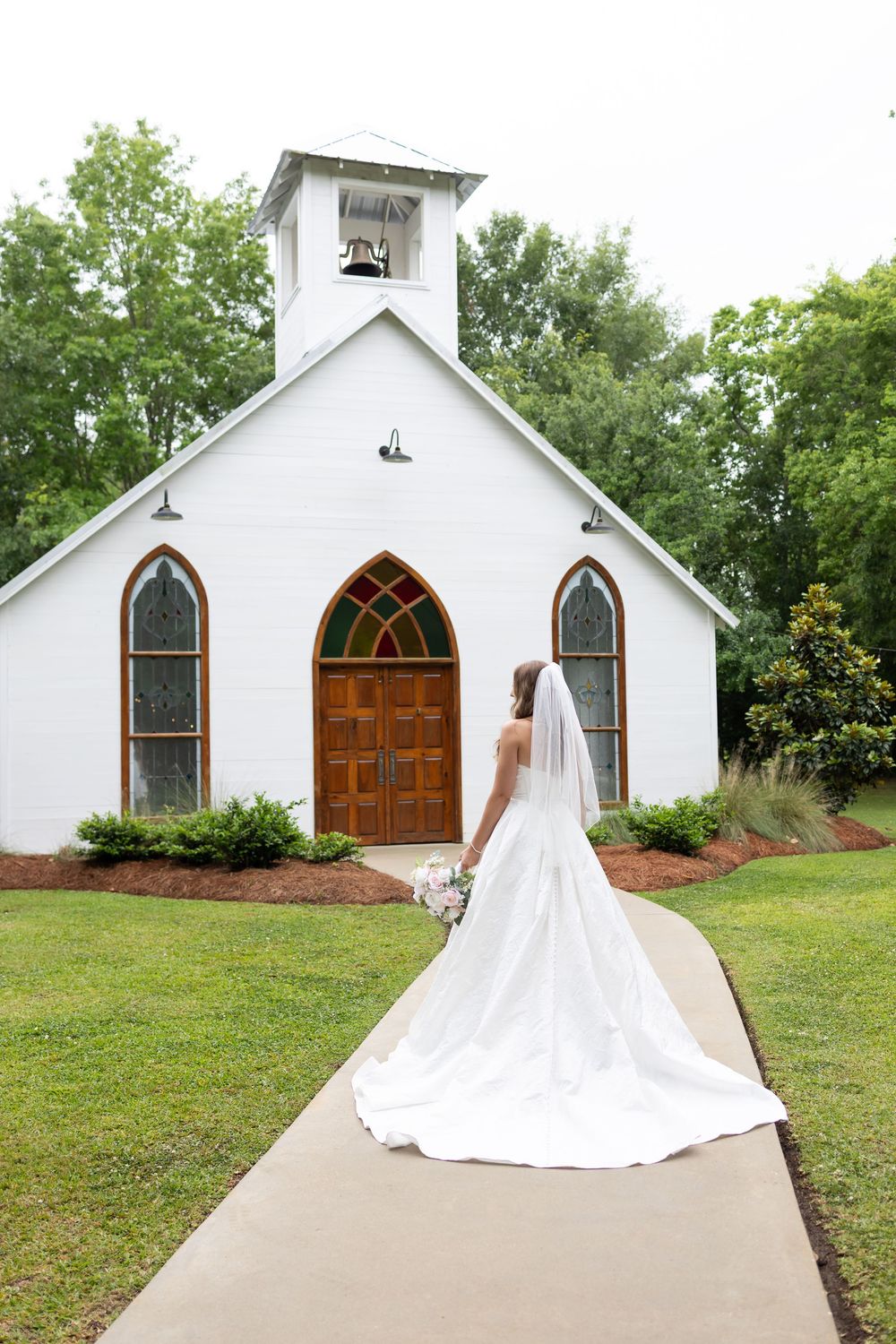 Bride in white gown and veil walks toward a charming white chapel with stained glass windows on her wedding day.