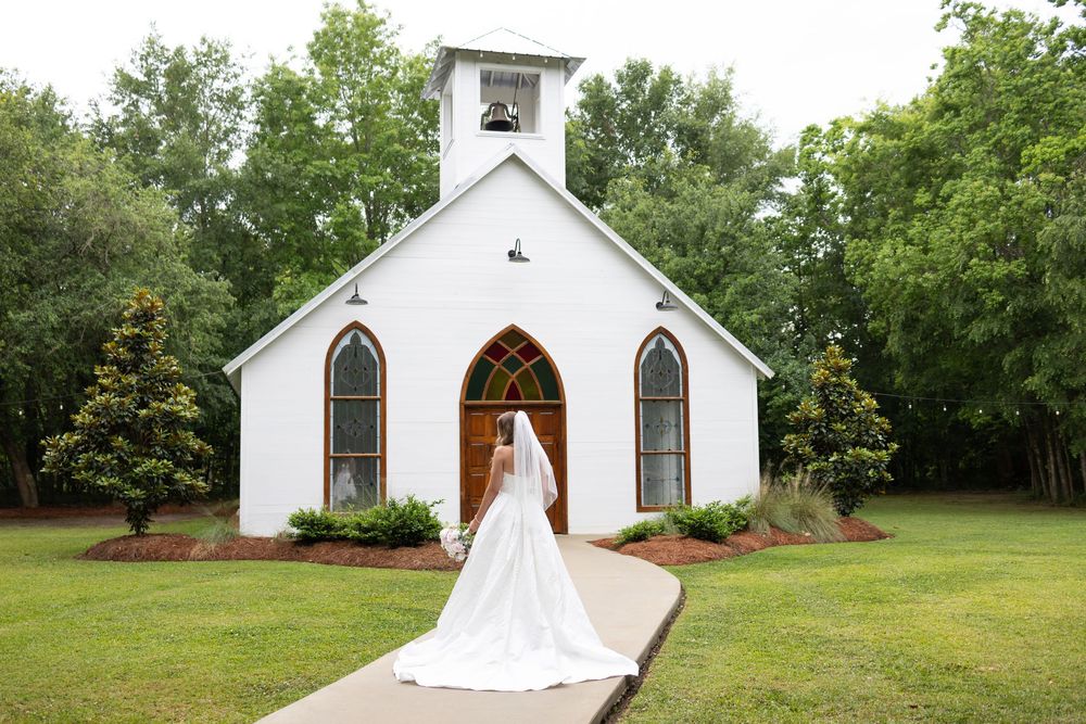 Bride in white gown and veil stands before a charming white chapel surrounded by lush green trees on her wedding day.