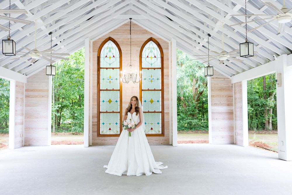 Bride in white gown stands before stained glass windows in an open-air chapel surrounded by lush greenery.