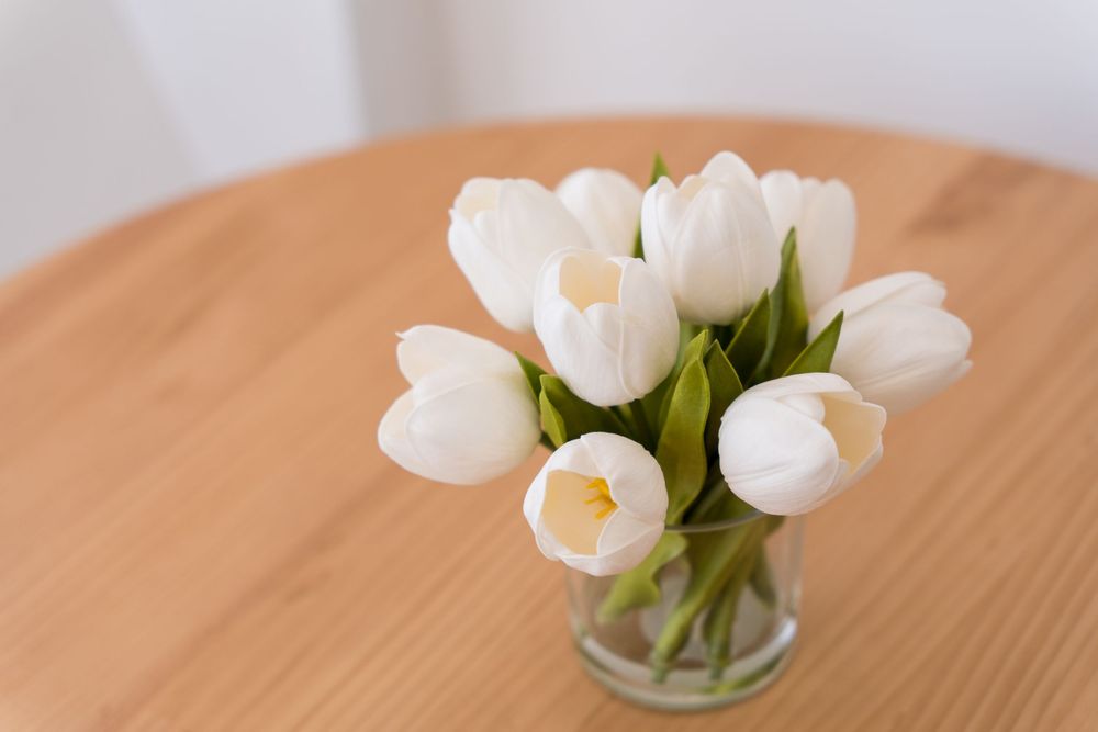 White tulips in a clear glass vase on a wooden table, soft natural light highlighting the fresh blooms.