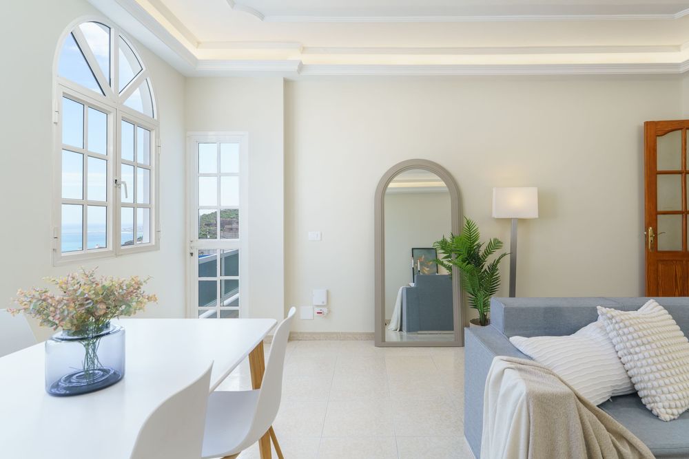 Bright modern living room with arched mirror, blue sofa, white dining set, and large arched window with sea view.