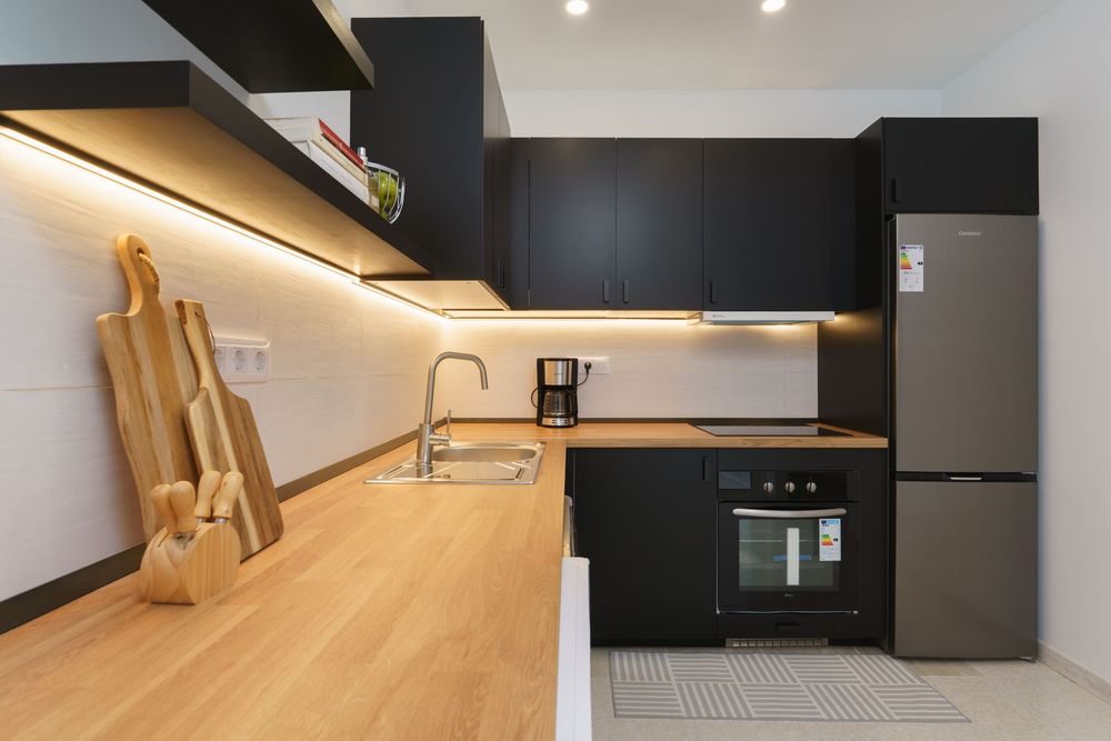 Modern kitchen with black cabinets, wooden countertop, stainless steel sink, built-in oven, and refrigerator with LED lighting.
