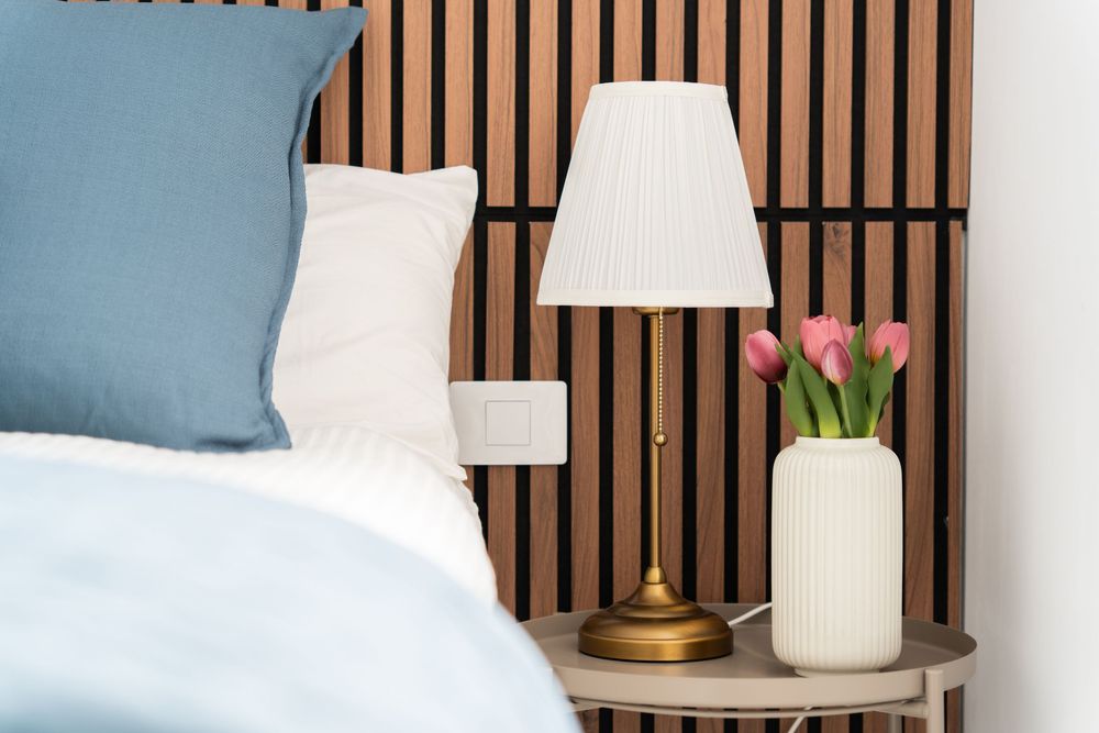 Cozy bedroom nightstand with brass lamp, pink tulips in white vase, blue pillows, and wooden slat headboard.