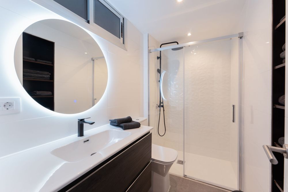 Modern bathroom with round LED mirror, floating dark vanity, white sink, and glass-enclosed shower with black fixtures.