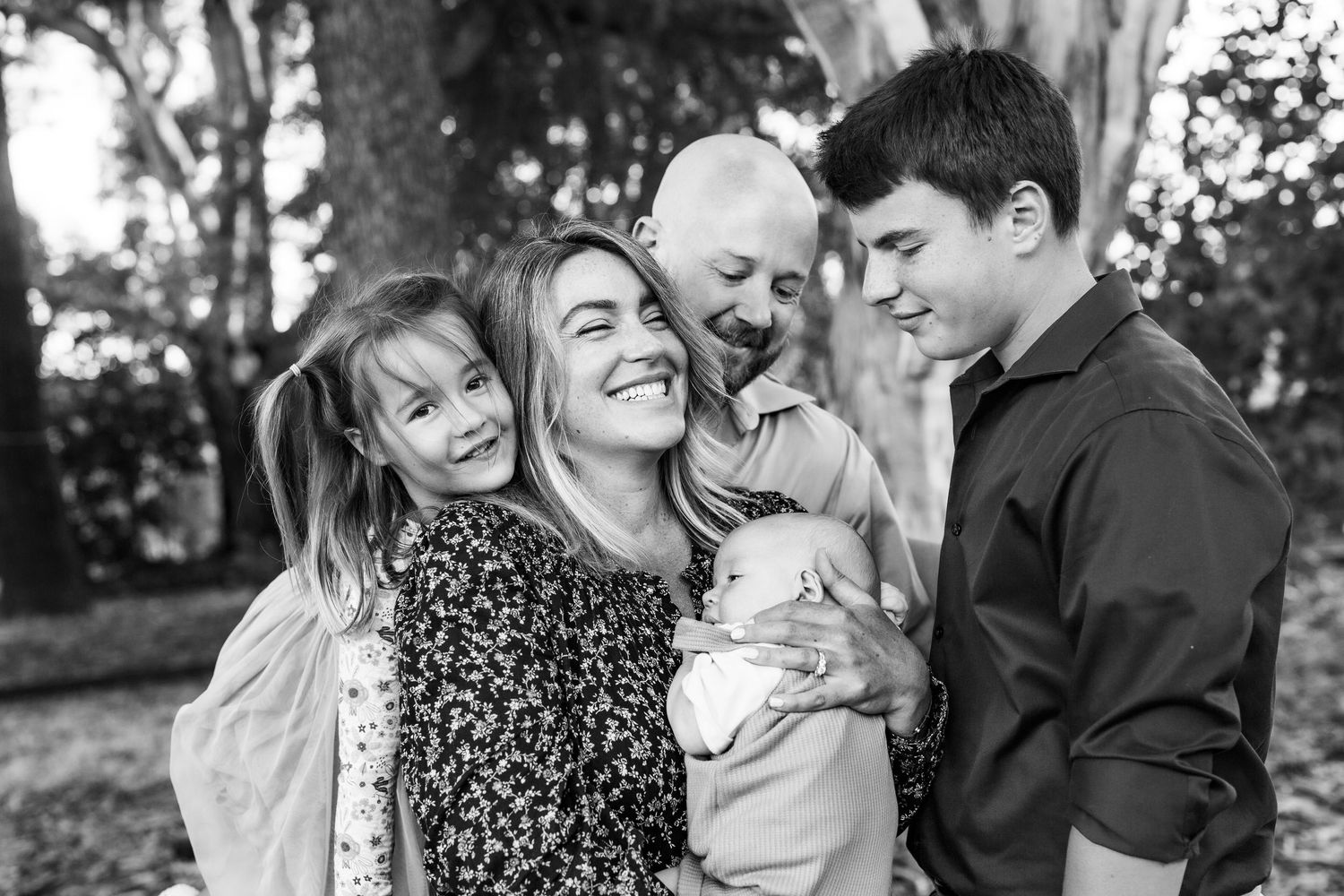 Black and white family portrait of parents with three children laughing and embracing outdoors near a tree.