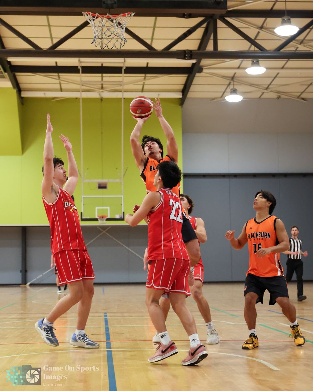 Basketball players compete in an indoor gym, with one player shooting over defenders in red uniforms during a game.