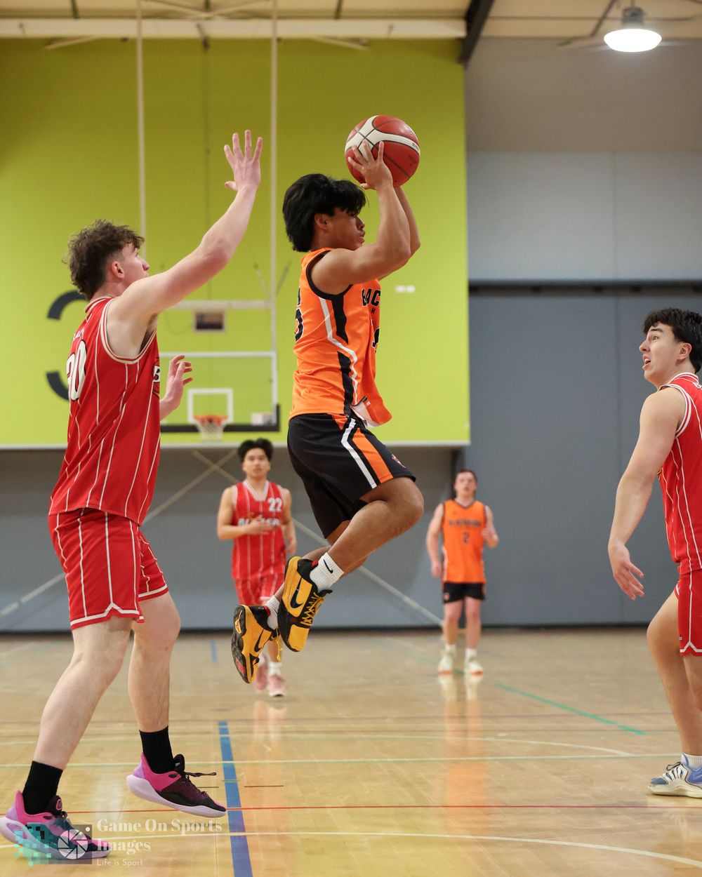 Basketball player in orange jersey jumps to shoot over defender in red during indoor youth basketball game.