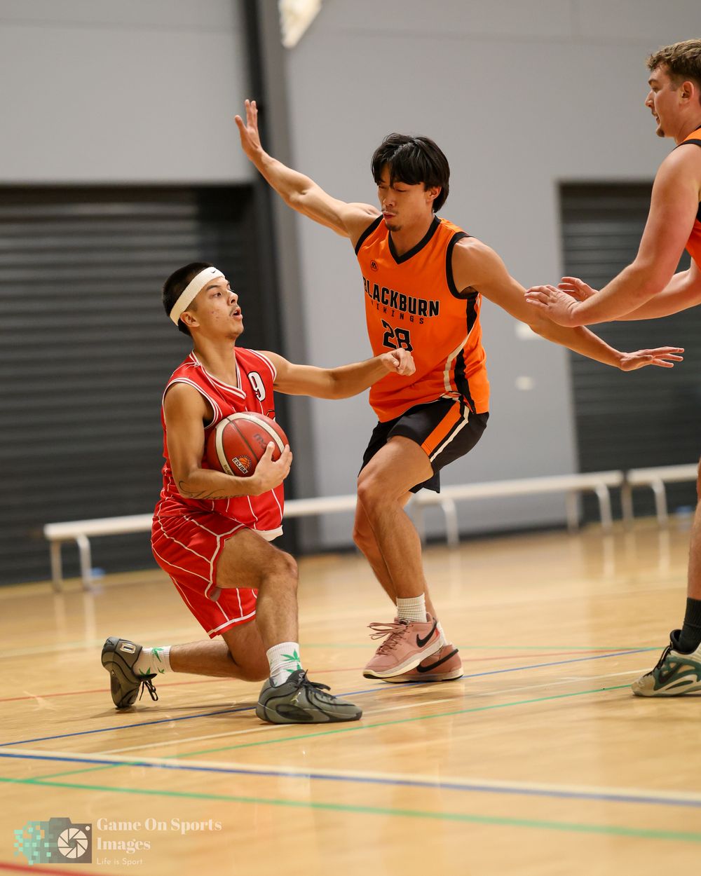 Basketball player in red kneels with ball while Blackburn Tigers defender in orange attempts a block indoors.