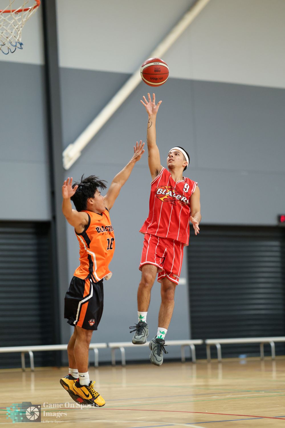 Two basketball players compete, one in red jumping to shoot over an orange-uniformed defender in an indoor court.