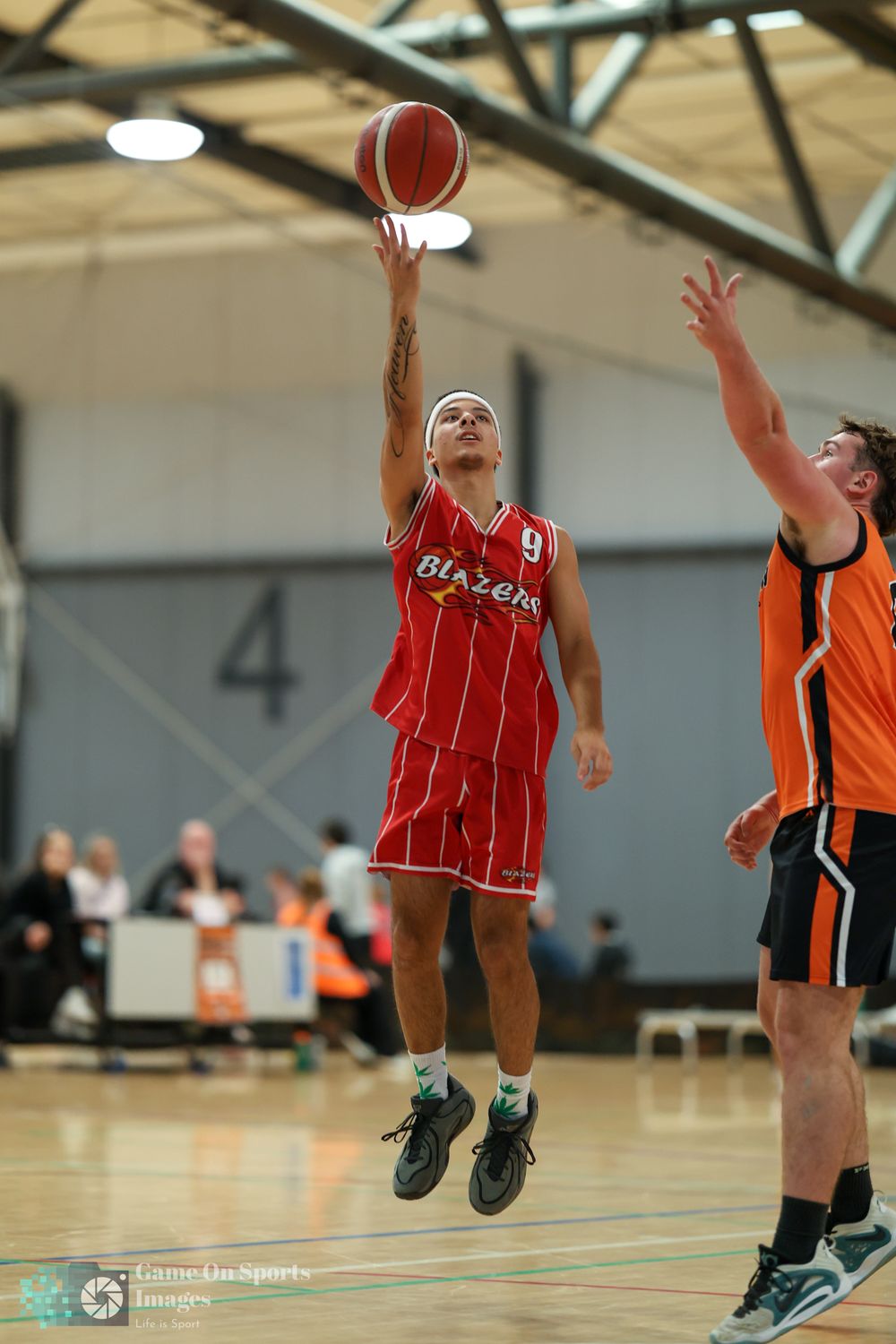 Basketball player in red Blazers jersey #9 leaps to shoot while opponent in orange uniform attempts to block.