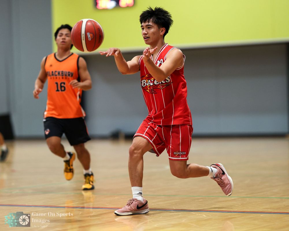 Young basketball player in red uniform dribbling ball during indoor game, opponent in orange Blackburn jersey visible.