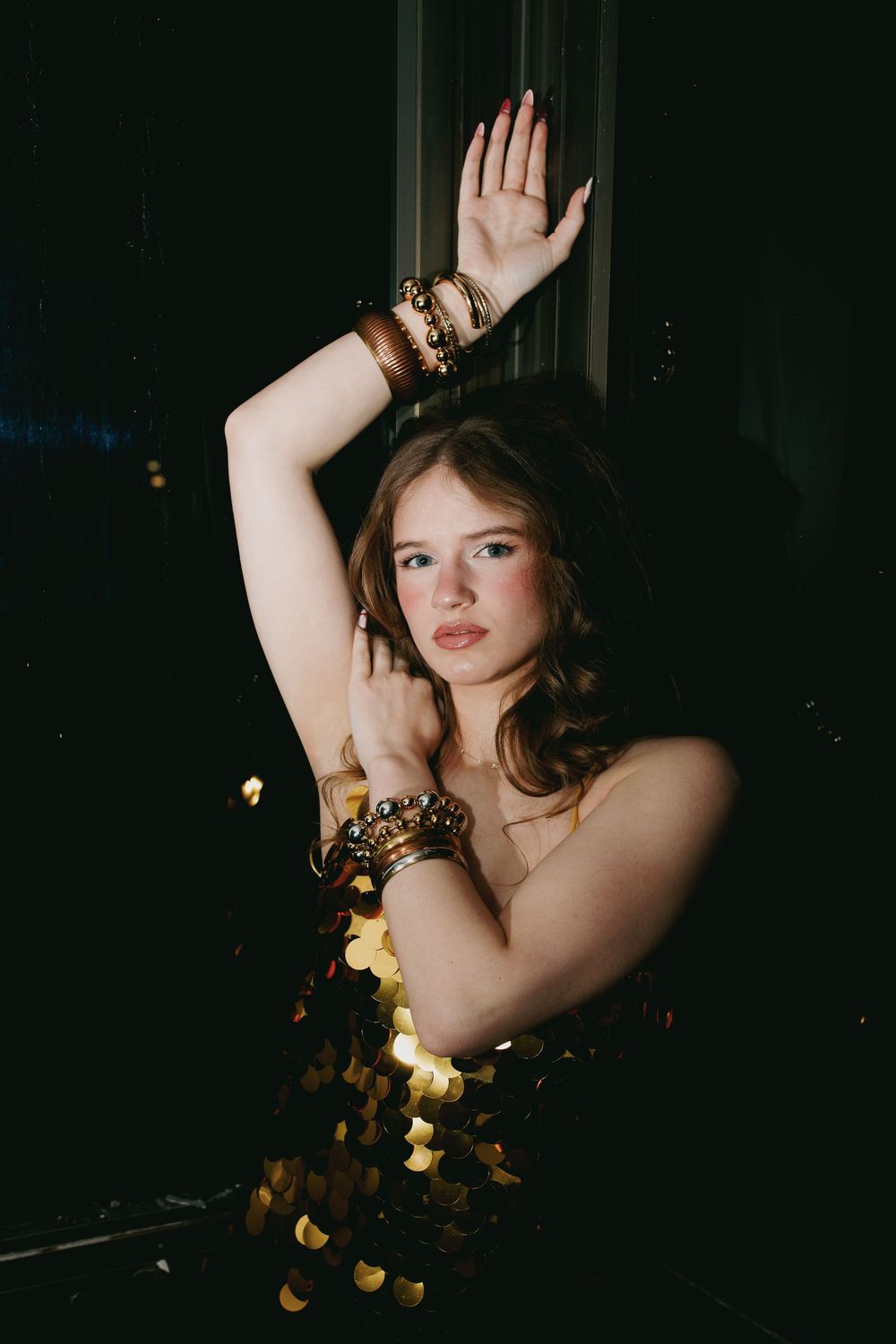 Young woman in gold sequin dress with stacked bracelets poses gracefully against dark background.