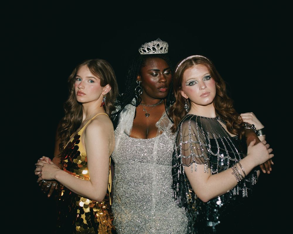 Three women in glamorous sequin gowns pose together, with the center woman wearing a sparkling tiara.