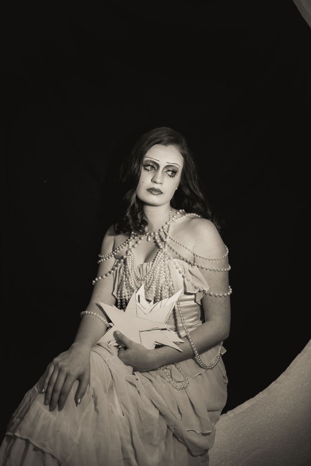 Black and white portrait of a woman in vintage dress adorned with pearl necklaces, seated against dark background.