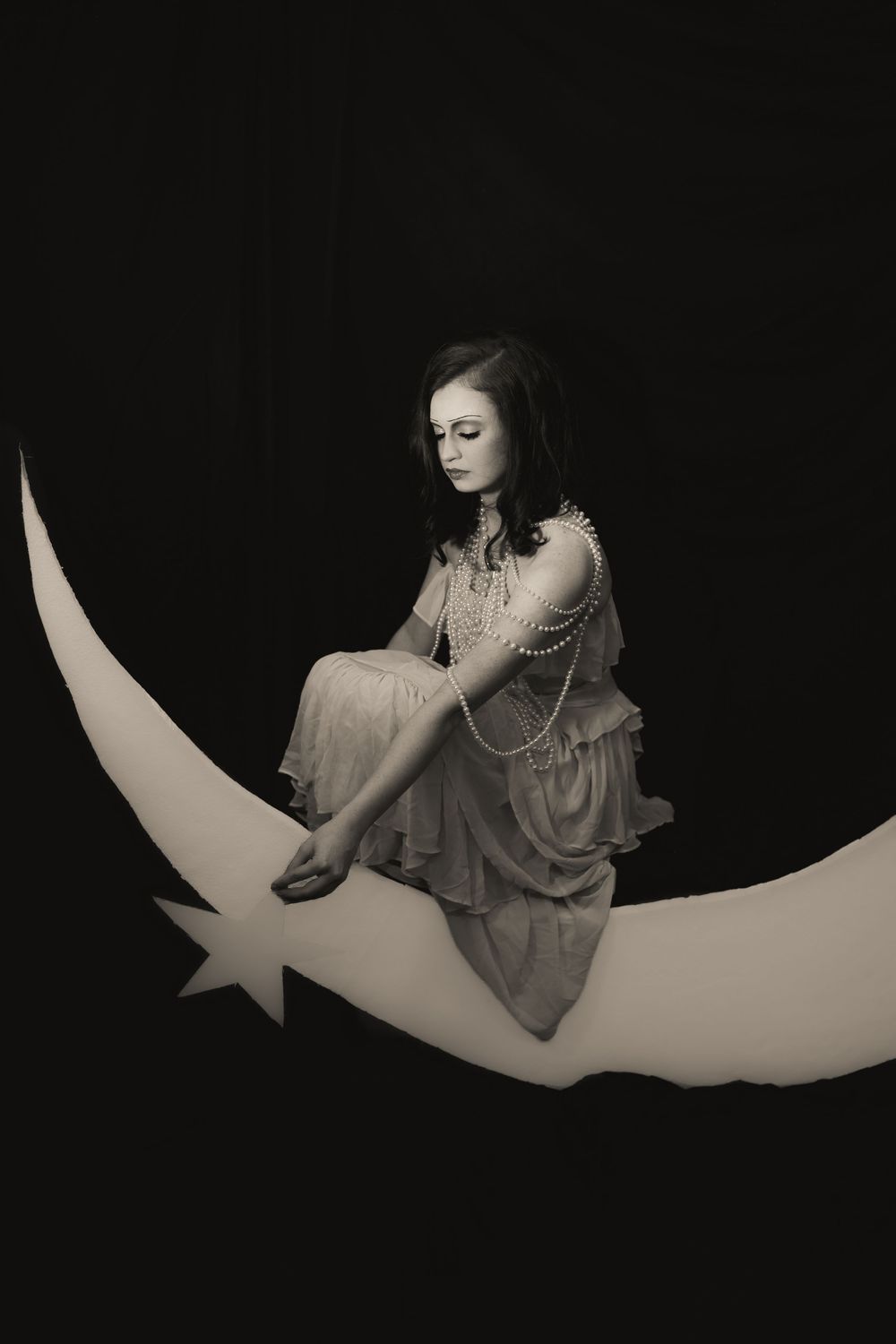 Black and white photo of a young woman in a vintage dress sitting on a crescent moon prop against a dark background.