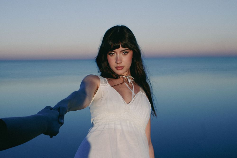 Young woman in white dress holding hands near calm blue ocean at dusk with gradient sky backdrop.