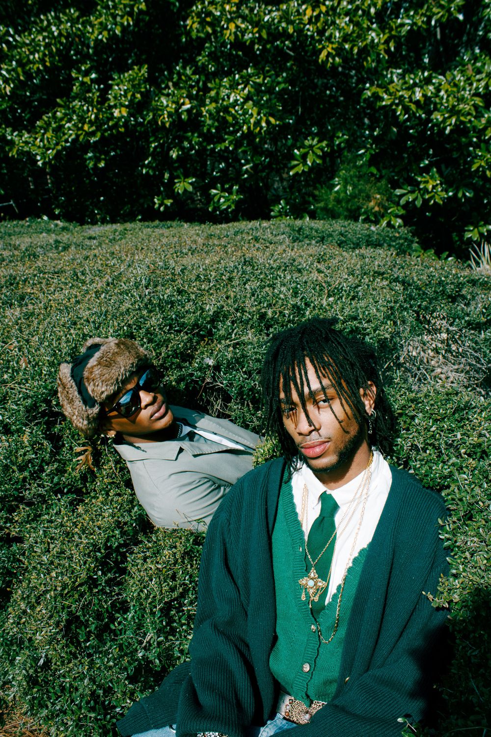 Two young men with dreadlocks relaxing on green hedges outdoors, one wearing sunglasses and a gray suit.