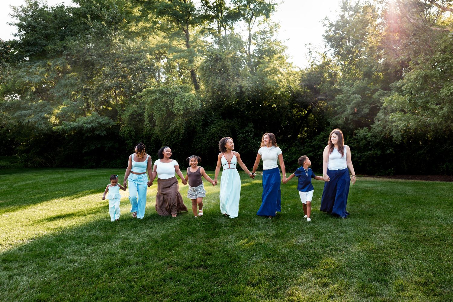 A group of women and children holding hands and walking together on green grass with sunlit trees in the background.
