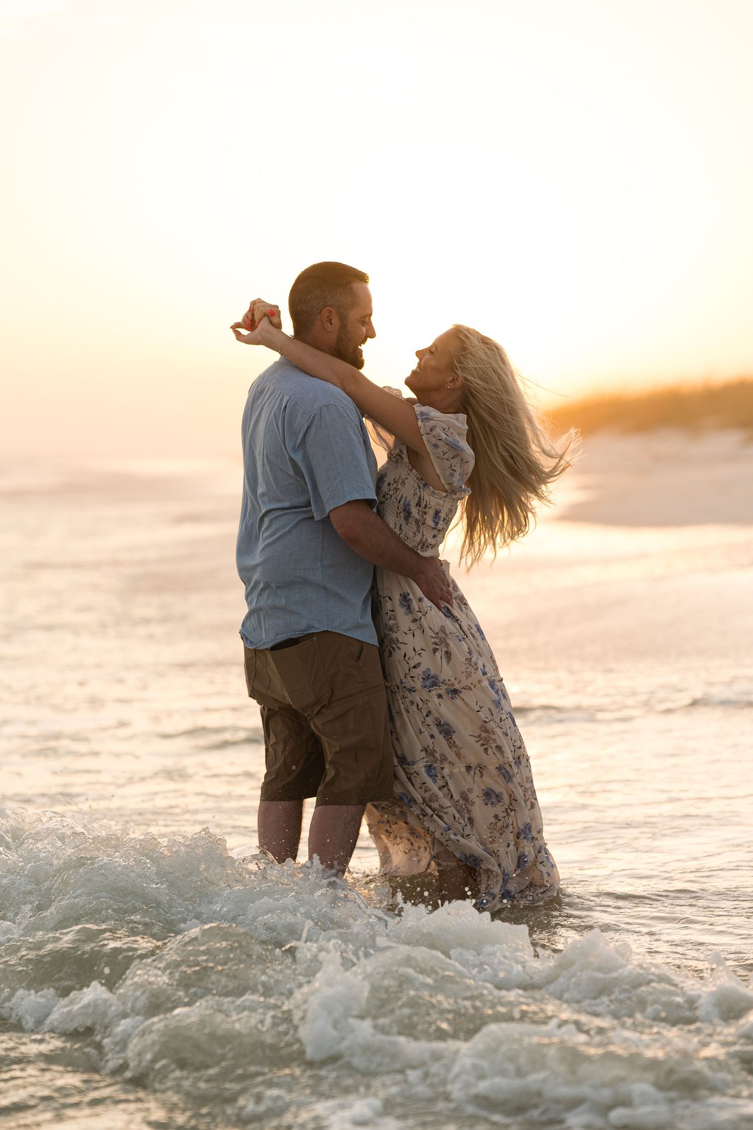 Romantic couple embracing in ocean waves at sunset on a beach, woman in floral dress, man in casual shorts.