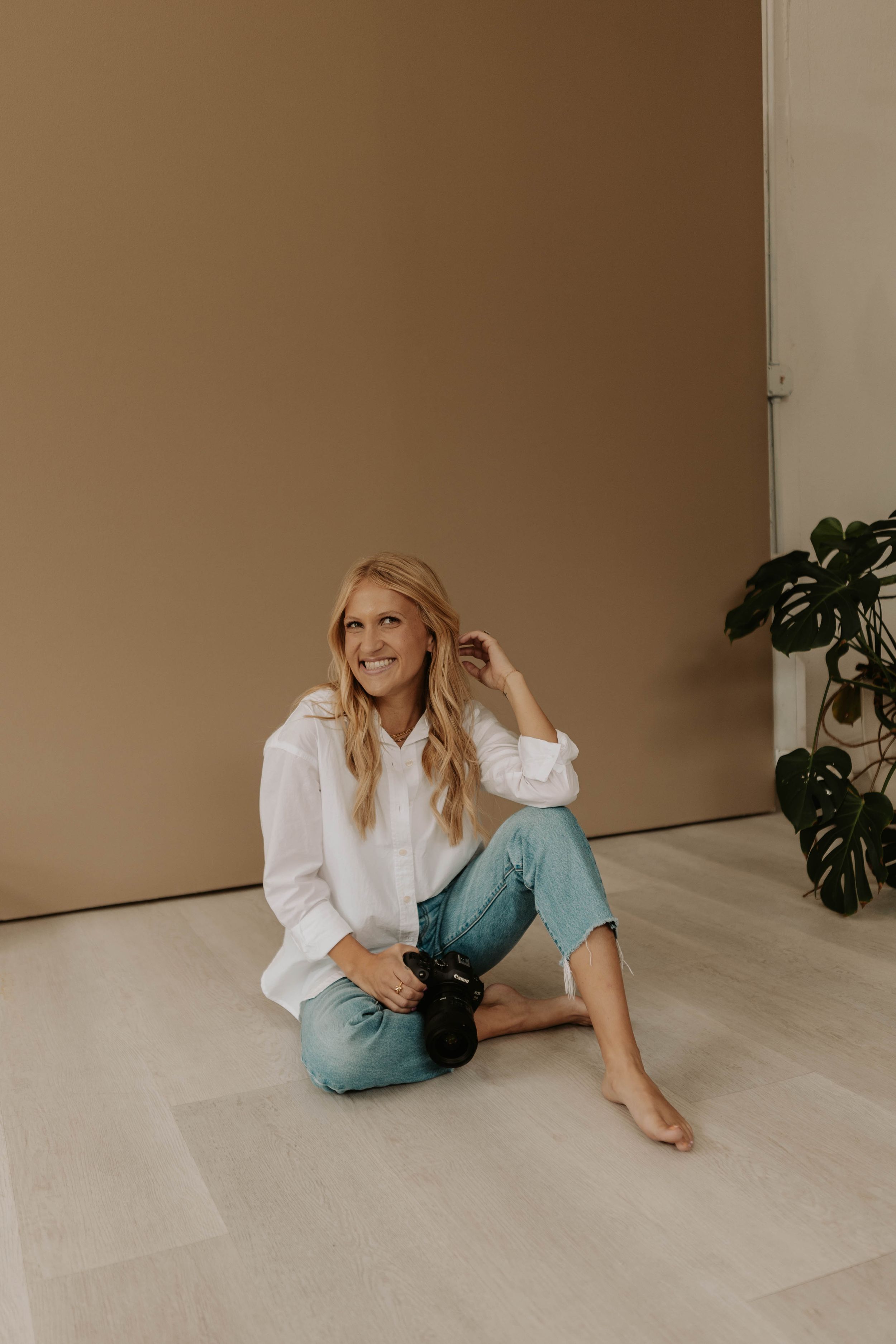 Smiling blonde woman sitting on floor in white shirt and jeans, holding camera, with plant and beige wall background.