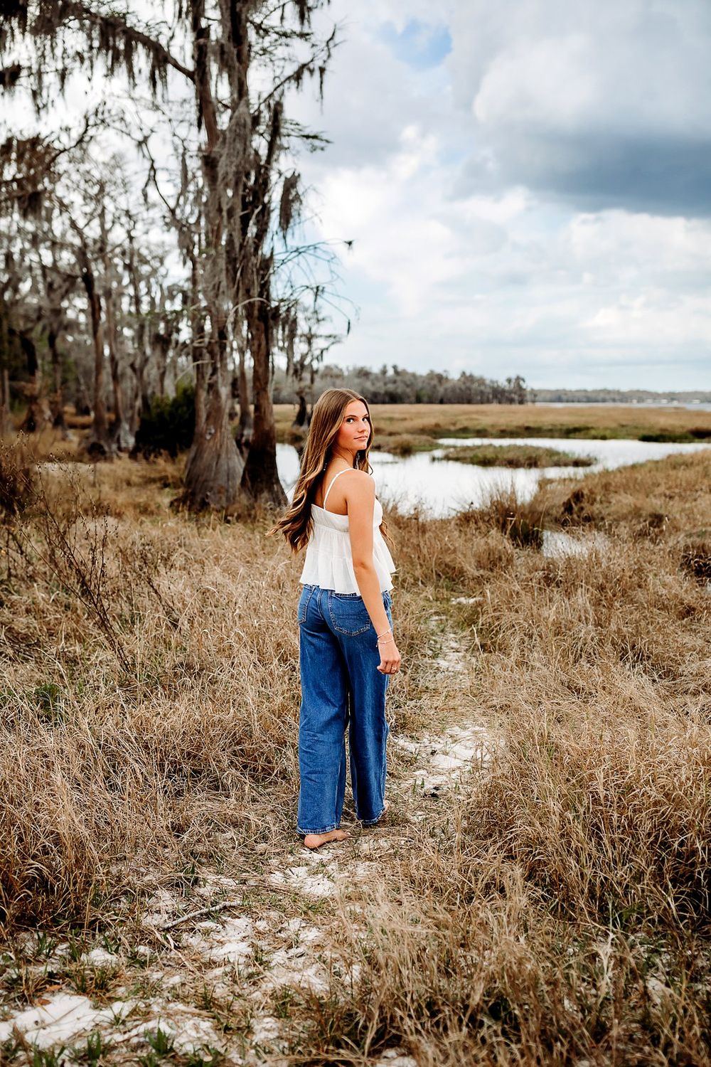 Young woman in white top and jeans standing in a coastal marsh landscape with Spanish moss-draped trees.