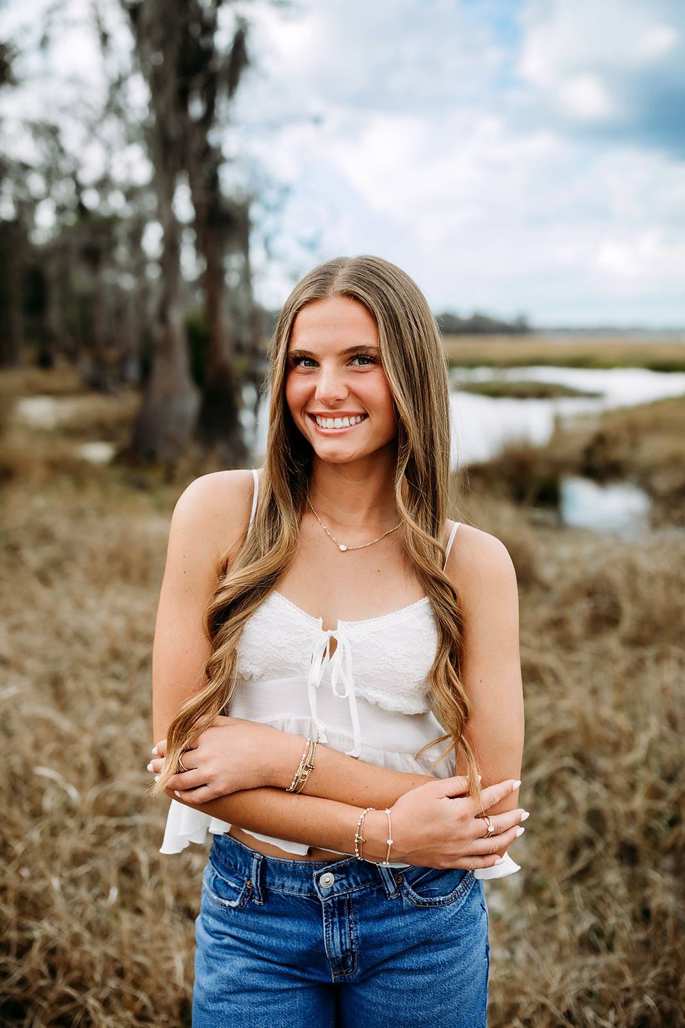 Smiling young woman in white crop top and jeans posing outdoors near a marsh with trees and cloudy sky.