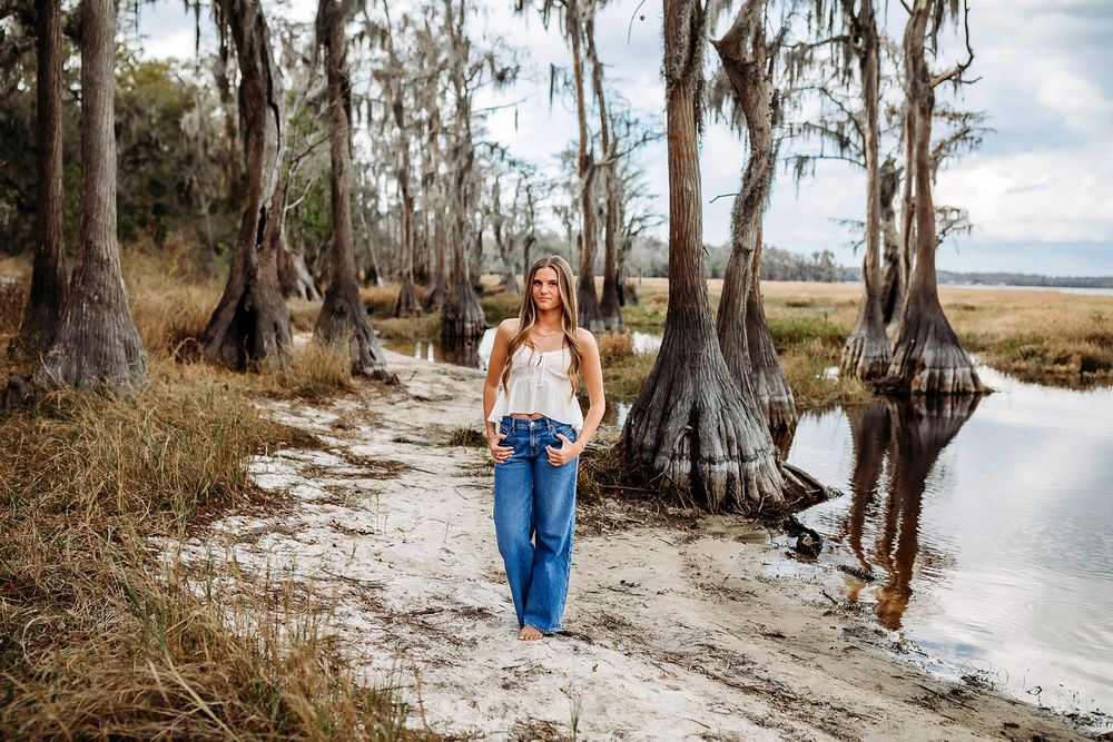 Woman walking on sandy path near cypress trees draped in Spanish moss beside a tranquil marsh waterway.