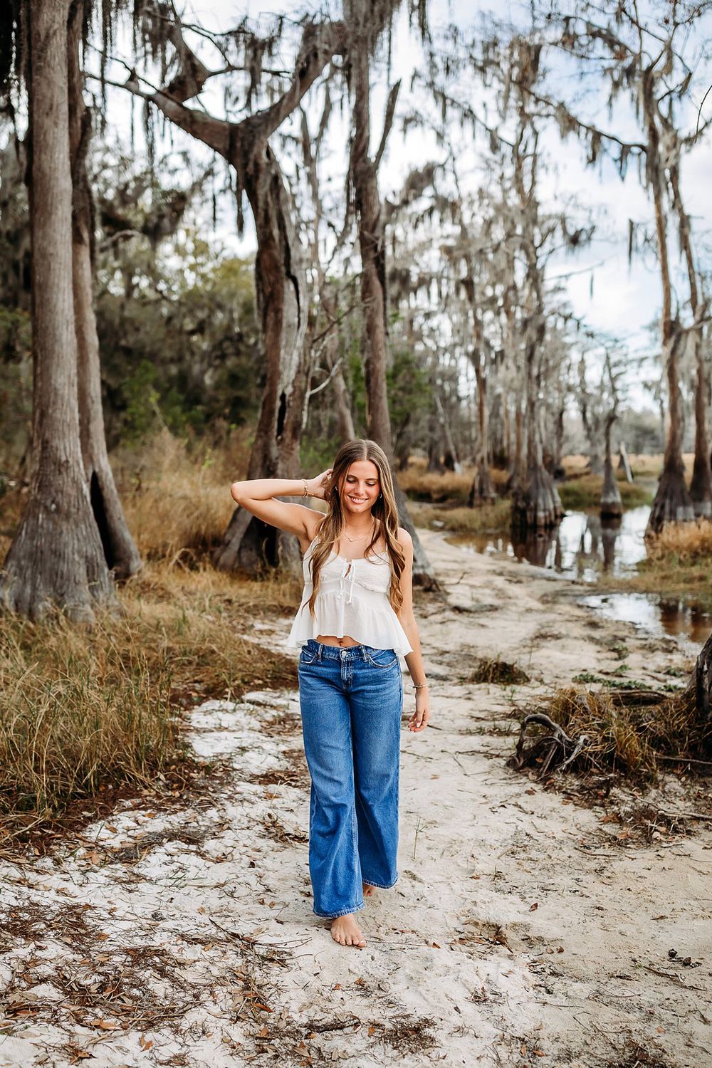 Woman in white top and wide-leg jeans walking barefoot on sandy path among moss-draped cypress trees near a swamp.