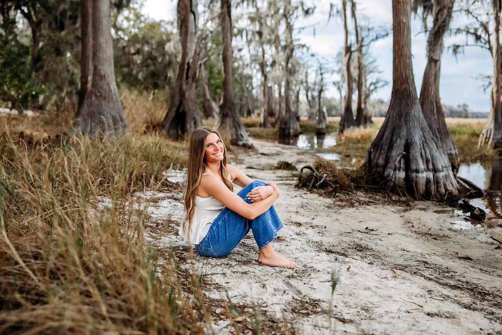 Young woman in blue jeans sits barefoot on sandy ground among cypress trees near a serene wetland.