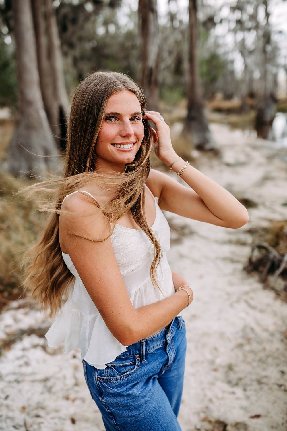 Smiling young woman with long hair in white top and jeans poses outdoors on sandy path among tall trees.