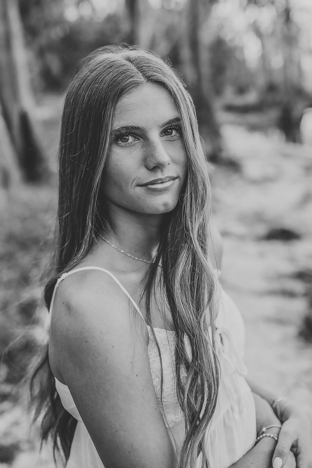 Black and white portrait of a young woman with long hair wearing a sundress, smiling softly in a forest setting.