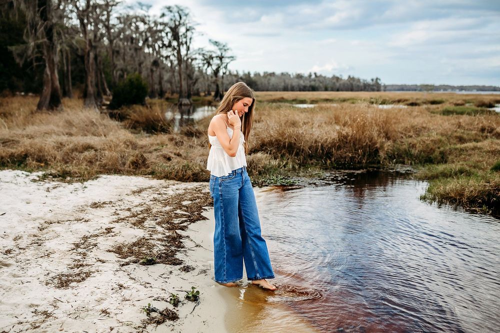 Young woman in white top and wide-leg jeans wading through a shallow creek in a coastal marsh landscape.